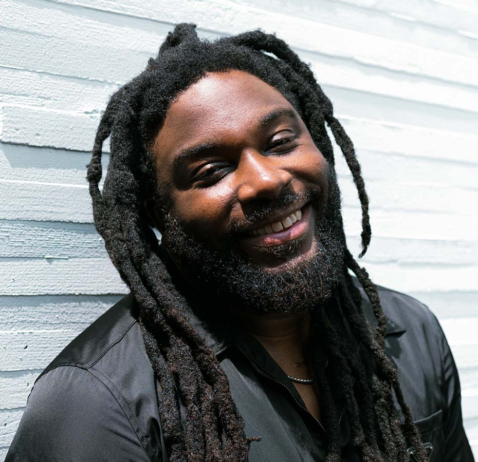 Author Jason Reynolds, a Black man with long dreadlocks and a beard wearing a black shirt and silver chain around his neck, is smiling and standing outside in front of a white concrete wall at the Kennedy Center REACH.
