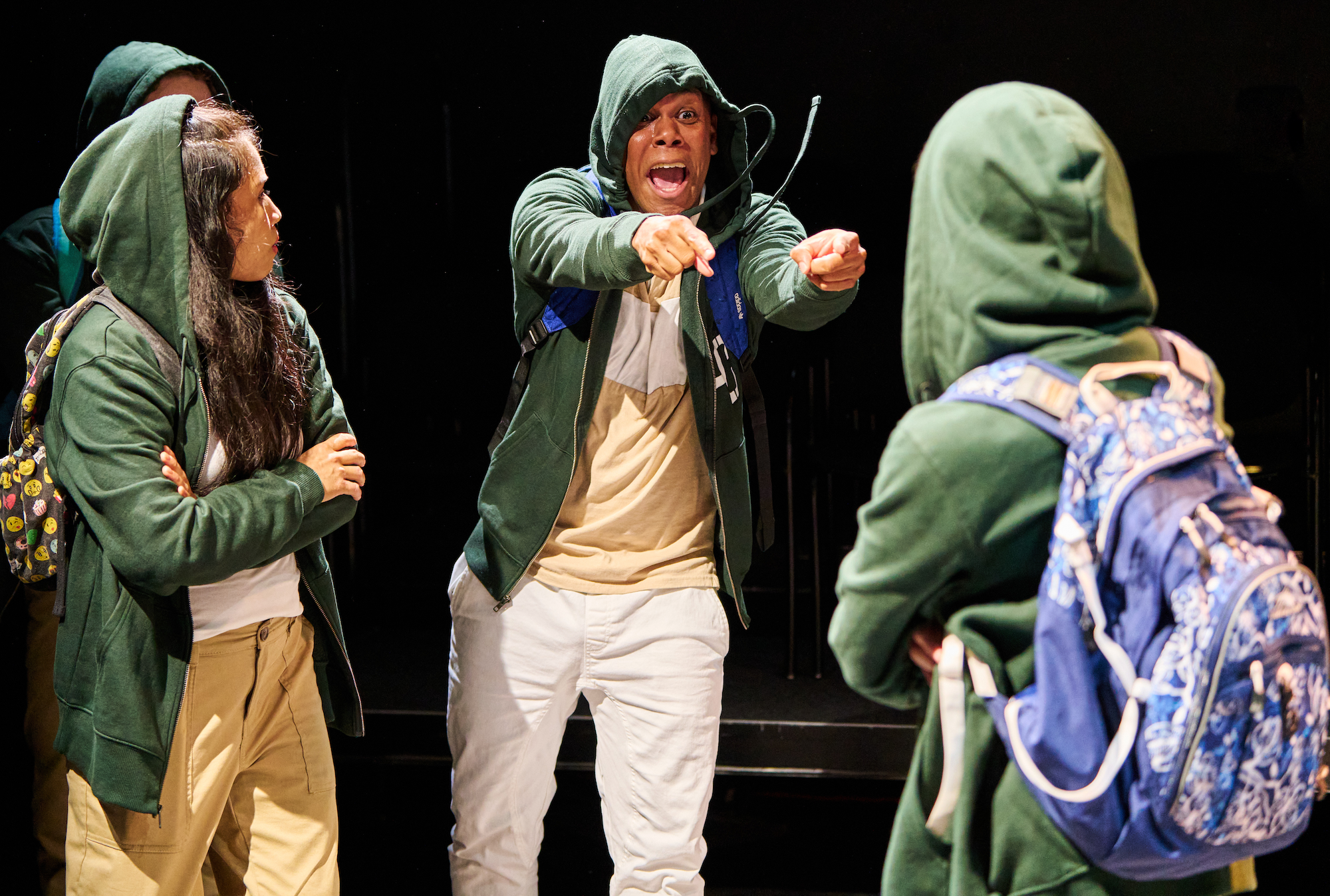 Four cast members on stage all wearing dark green hoodies and backpacks. One cast member is pointing at another with both hands and has a serious and upset look on his face. Another cast member with long hair and arms folded looks on.