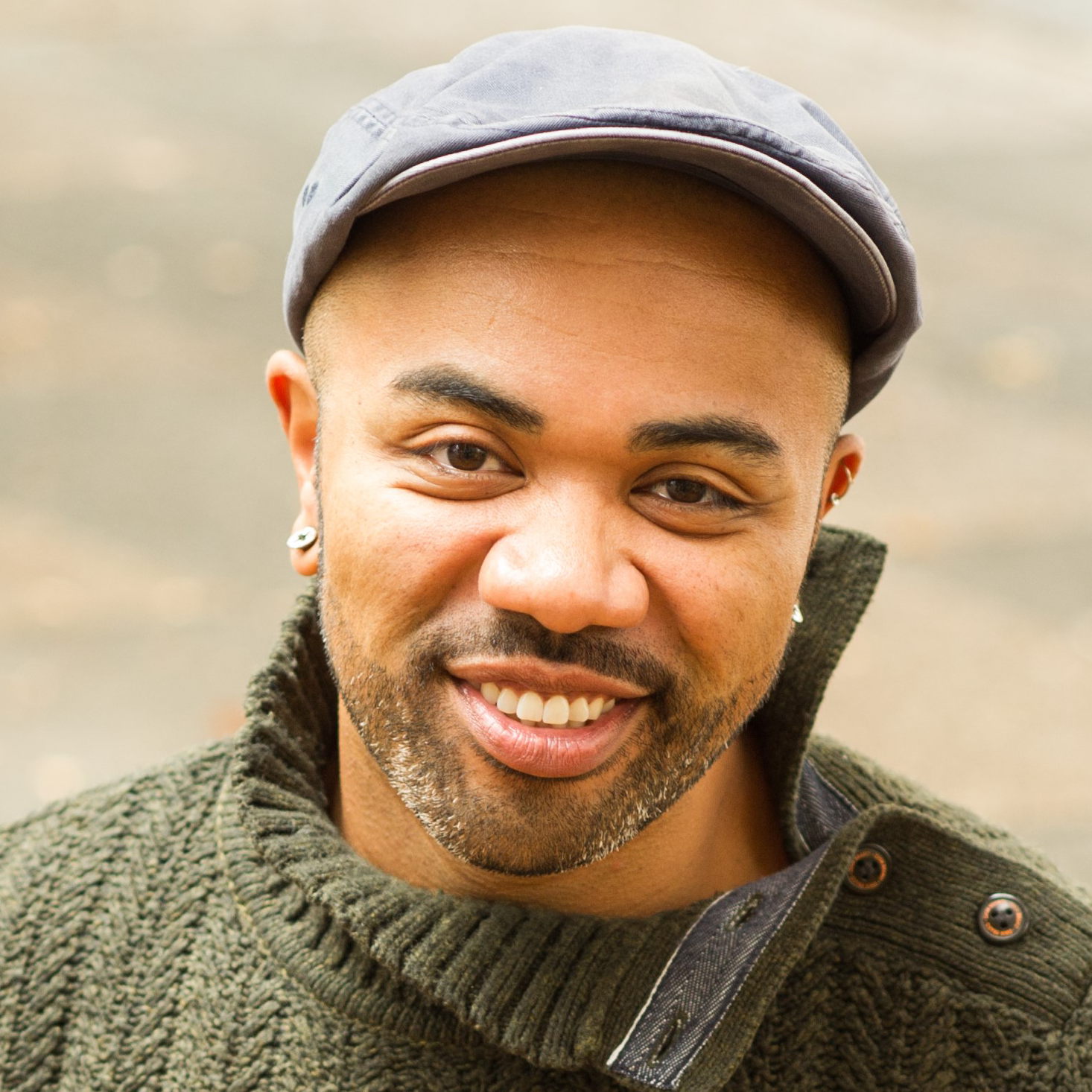 Headshot of Director Raymond O. Caldwell, a Black man with a short-trimmed graying beard wearing a green sweater and gray ivy cap smiling.