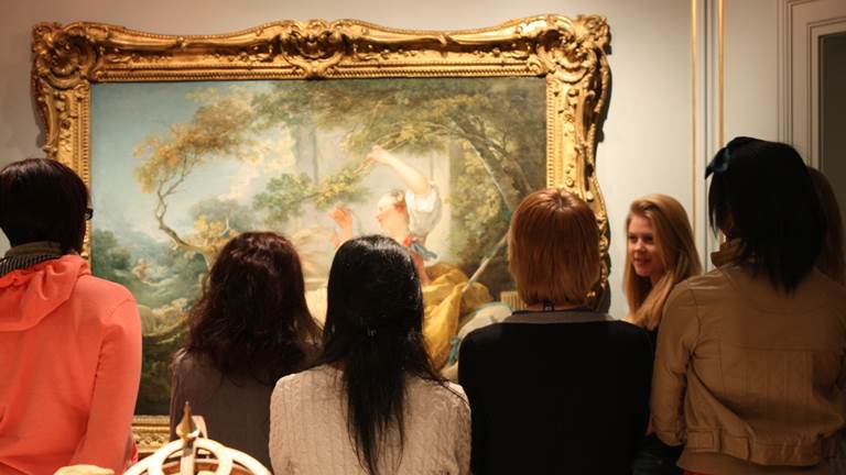 A group of students stand in front of a painting and listen to a guide 