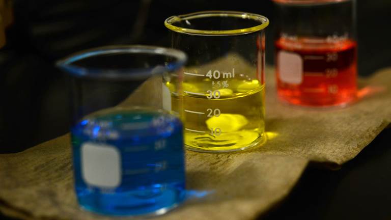 Three beakers filled with blue, yellow, and red liquid respectively sit side by side on a table