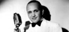 A black-and-white photo of jazz bandleader, arranger, and pianist Fletcher Henderson. He wears a white suit with a black bowtie and holds the stand to an old school microphone.