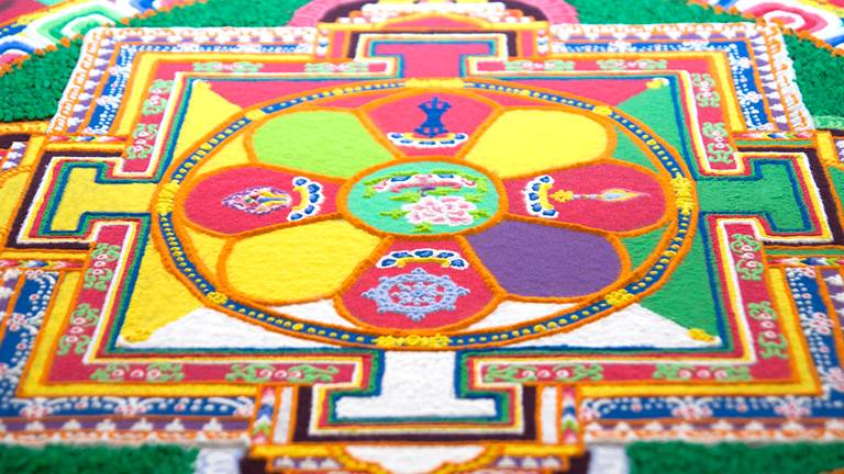 A sand mandala with a circle at the center and colorful patters around it.  