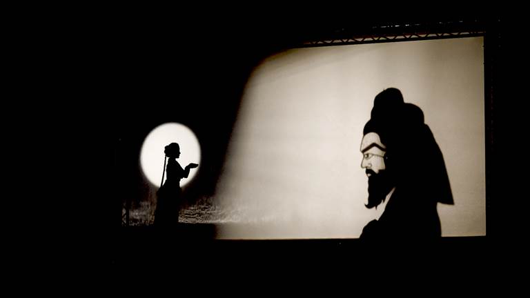 A woman stands on the left with her hands cupped. To the right is a shadow of a man’s head behind a large white screen. The man has a long beard and raised eyebrows that stretch across the side of his face. 