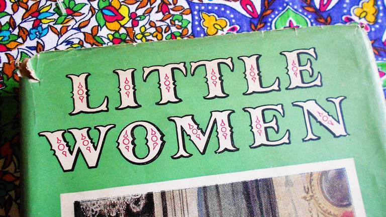 A book titled, "Little Women." The book cover is green and the title is written in white block letters, with black shadowing, small red decorations on the inside of the letters. The book sits on a floral backdrop.