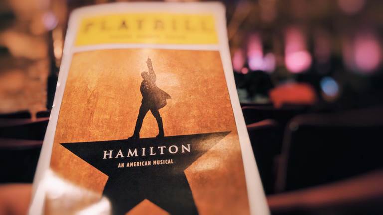 A Playbill book. A black star with four points and the silhouette of a man standing up with one hand pointing into the air. His body resembles the fifth and top point of the star. In the middle of the star there is white text that says, "Hamilton, An American Musical."