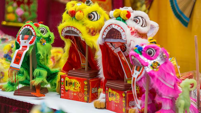 Four Chinese Lion Puppets on display. There are two small puppets, one is green and the other is pink. In the middle are two larger puppets, one is yellow and the other is red. 