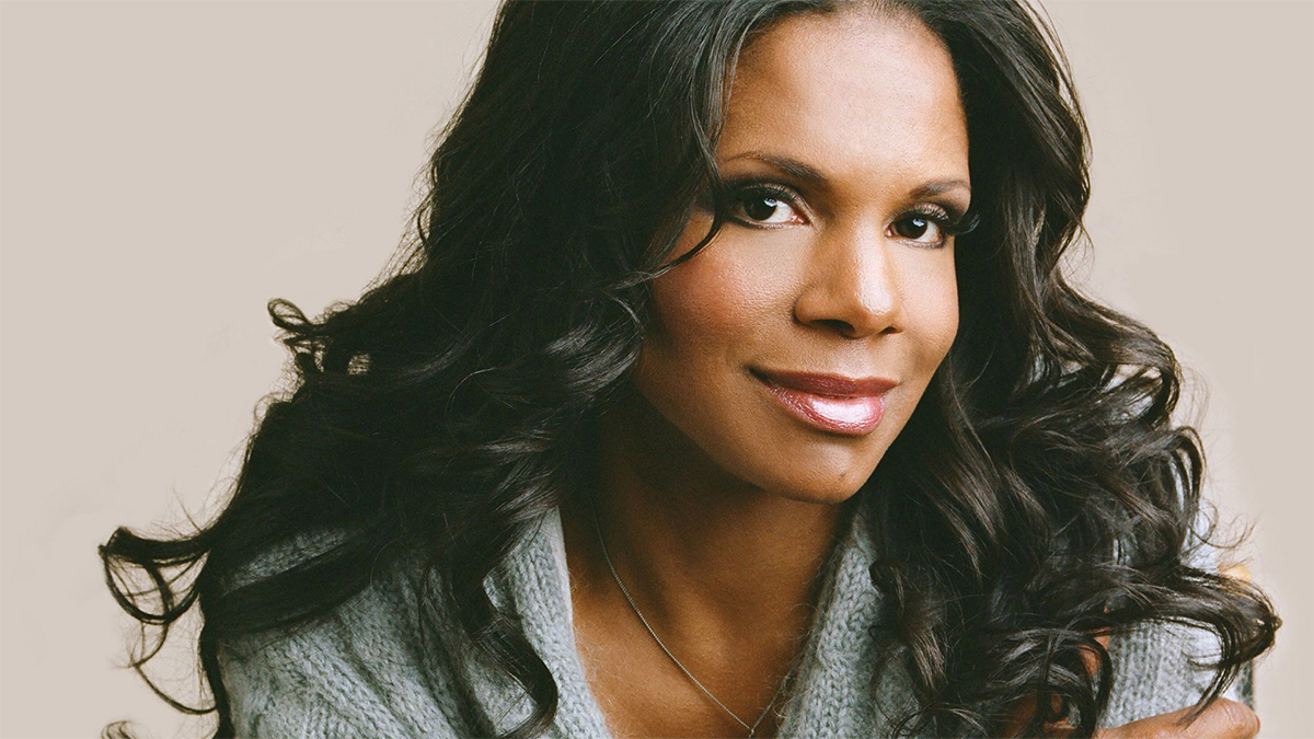 A head shot of singer Audra McDonald in front of a tan background. She wears a light gray sweater with a necklace, and her hair frames her face in soft wavy curls.