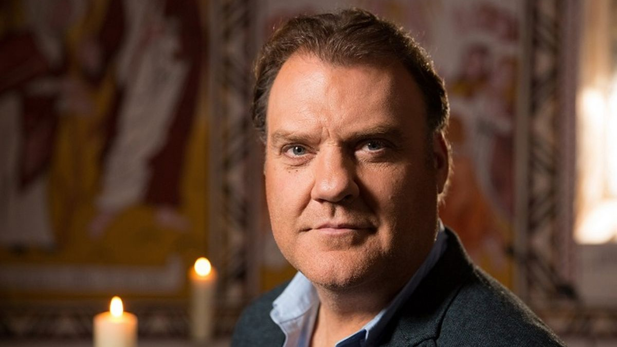 A head shot of singer Bryn Terfel in front of a decorated wall and two lit candles. He wears a dark jacket over a light blue dress shirt.