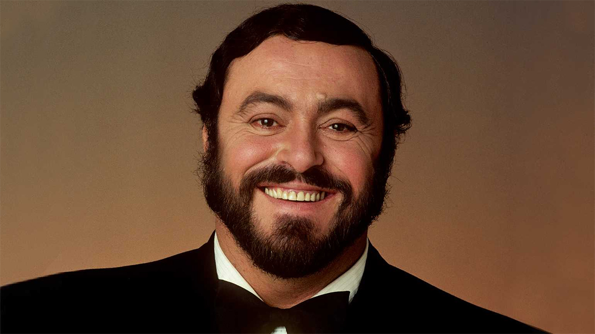A head shot of opera singer Luciano Pavarotti in front of a brown ombre background. He wears a black tuxedo jacket, black bowtie, and white dress shirt. He has a mustache and beard.