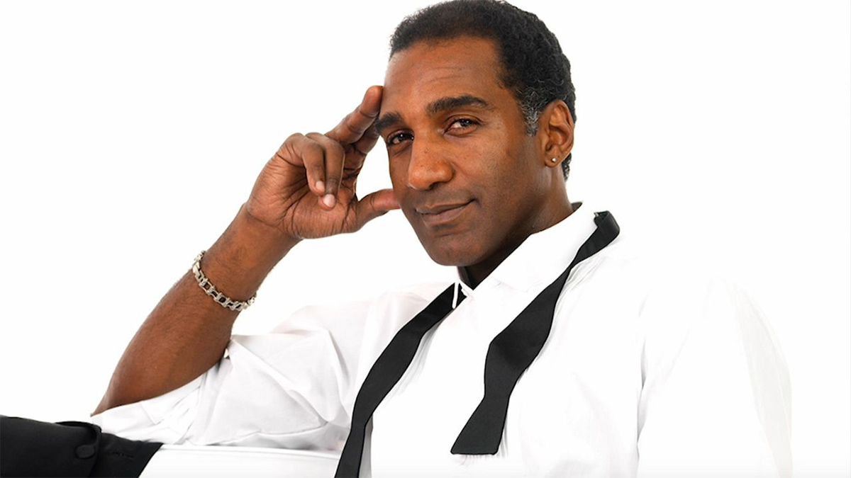 A photo of singer Norm Lewis in a white room, lounging on a white chair. He wears a white dress shirt with an undone black bowtie around his neck, a silver bracelet, and silver ear studs. His tuxedo jacket is draped over the chair.