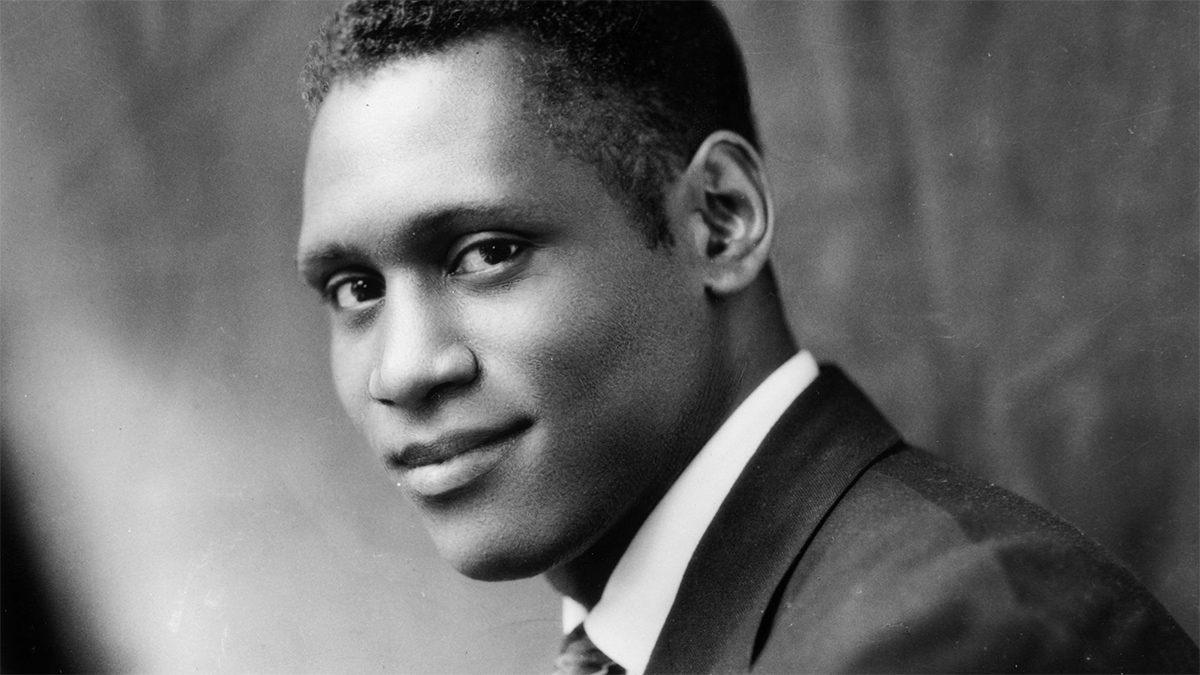 A black and white image of singer Paul Robeson smiling at the camera. He wears a dark jacket and tie with a white dress shirt.