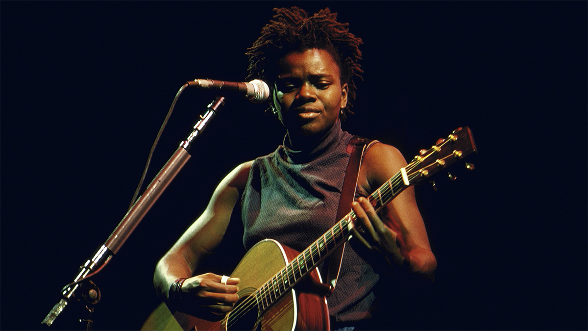 A photo of singer Tracy Chapman in front of a microphone while playing an acoustic guitar. She wears a dark sleeveless turtleneck top and a thumb ring, and her hair is in twists. She is under a spotlight in a dark space.