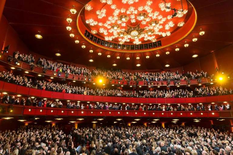The Kennedy Center Opera House with elaborate chandelier on the ceiling and a full house applauding an honoree in the Presidential Box.