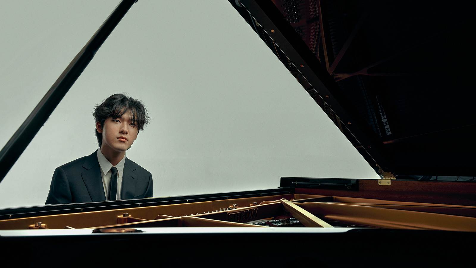 Artist, Yunchan Lim, sitting in front of a open grand piano. He has black hair and wearing a grey suit.