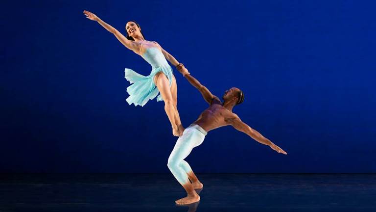Two dancers - a man holds hands with a woman who is standing on his knees as they both gesture outward on a blue background.