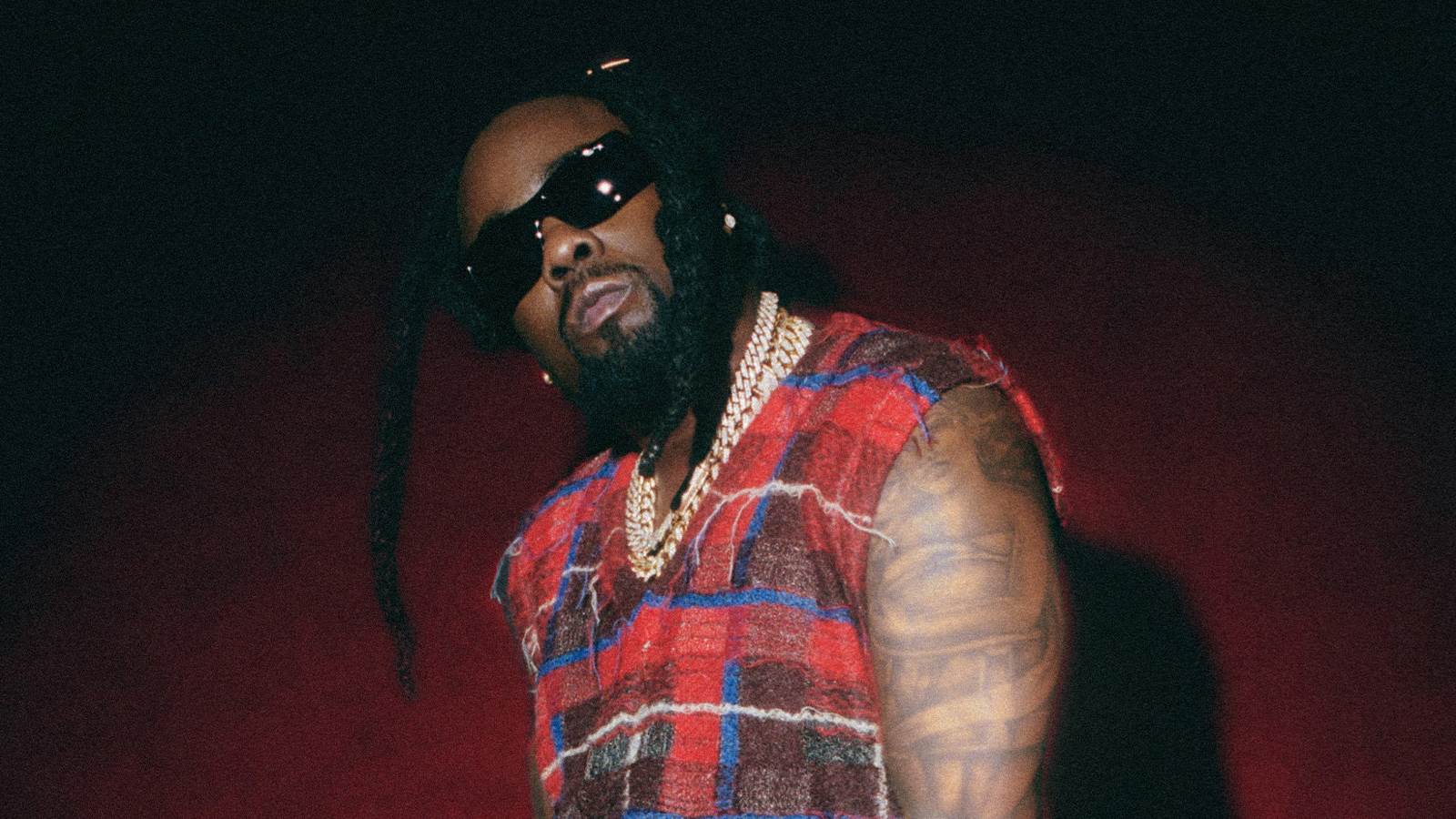 Wale stands against a dark red wall with black sunglasses and a plaid sweater vest. His arms are tattooed and he is wearing diamond earrings and gold chains. 