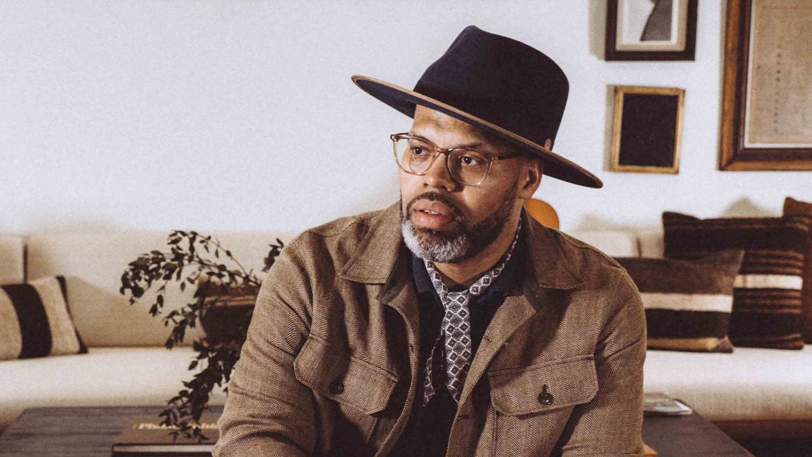 Eric Roberson is wearing a large, brown, brimmed hat and clear glasses. He looks off to the side and is sitting infront of a wood coffee table and white couch with pillows. 