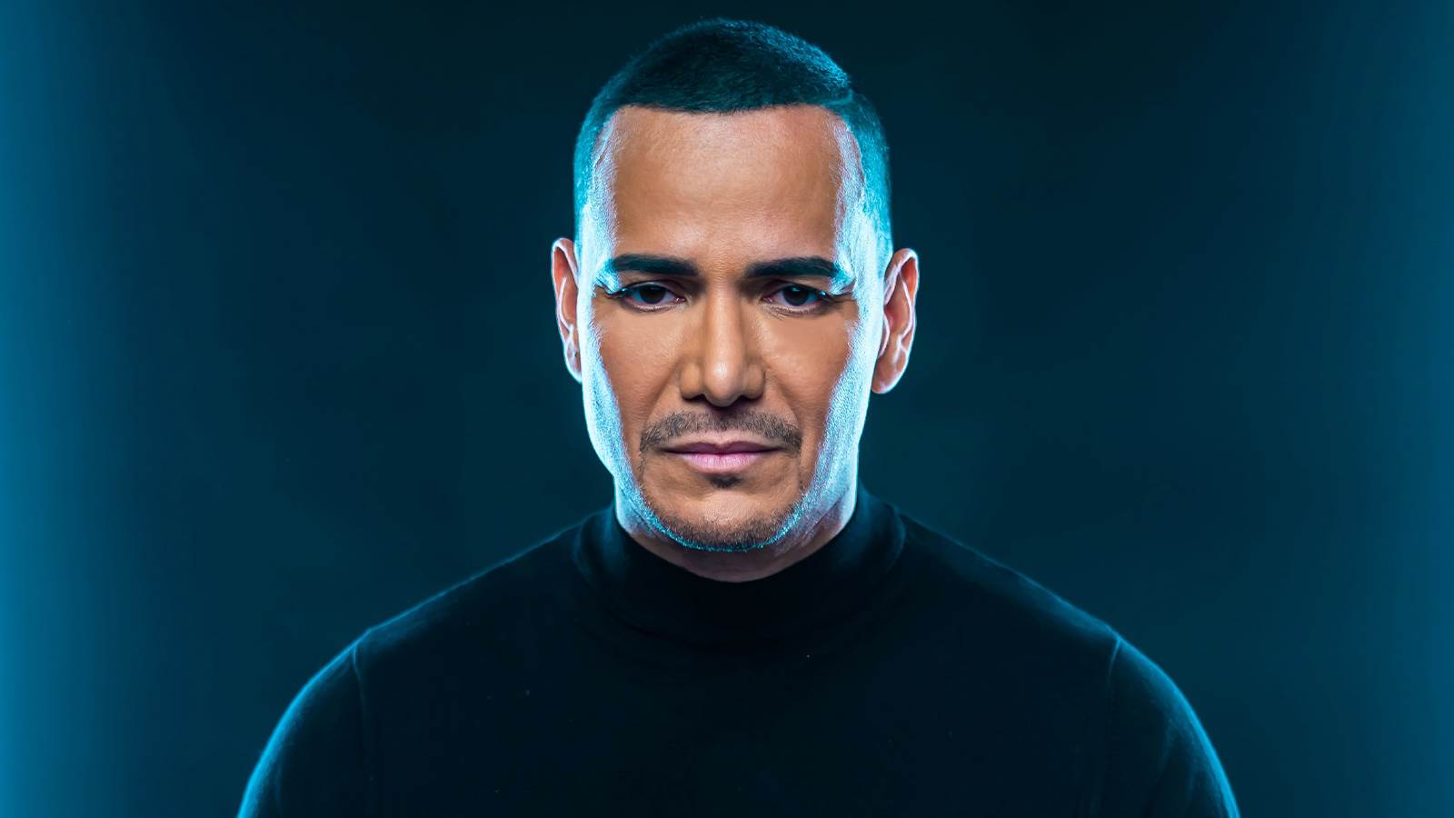 A close up of Victor Manuelle standing against a blue wall with a blue light on his face. 