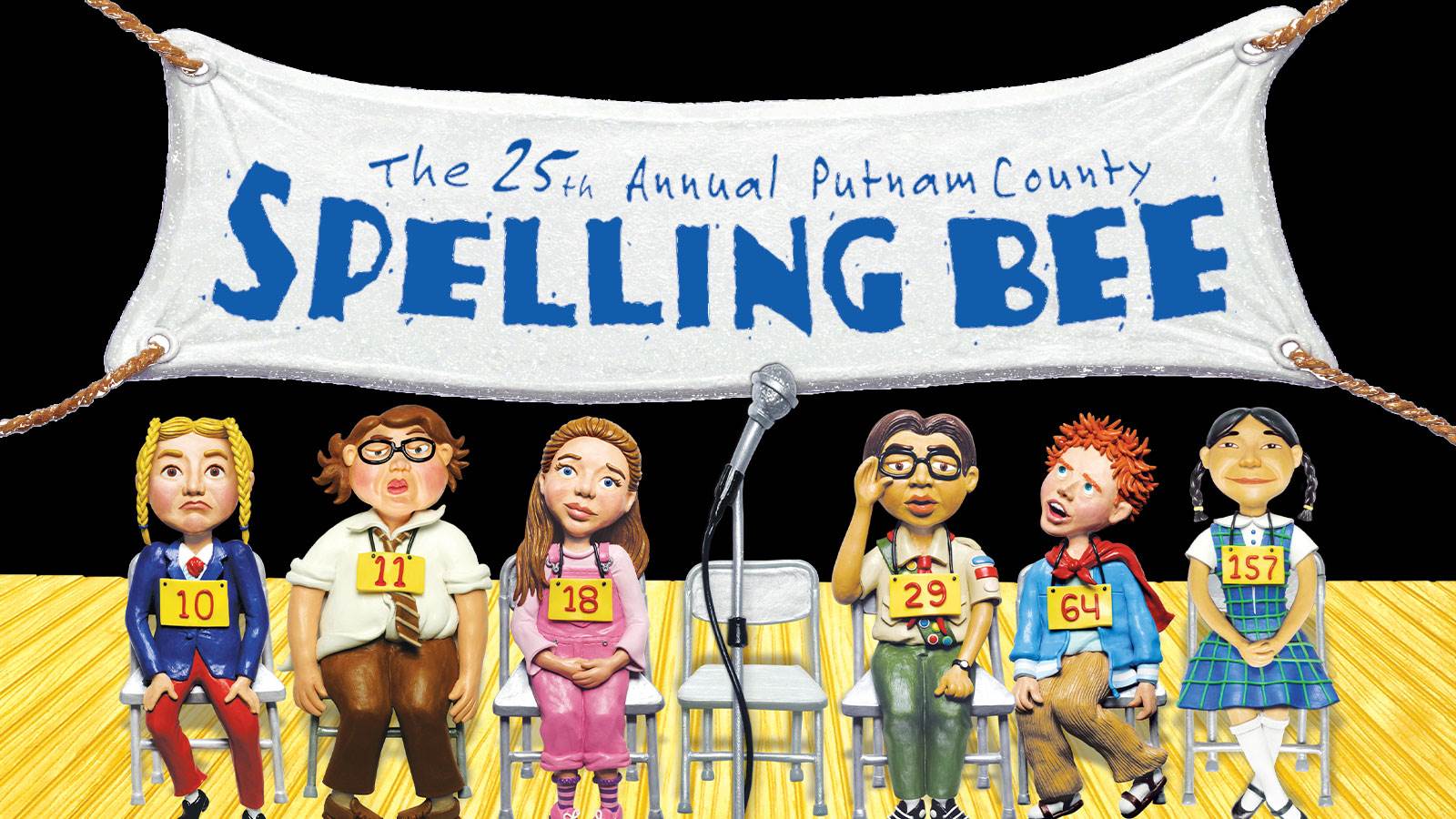 A white handmade banner is hung using rope at the top of the photo. It says "The 25th Annual Putnam County Spelling Bee". Below the banner is six children sitting on metal folding chairs and numbers pinned to their shirt. In front of them is a microphone. 