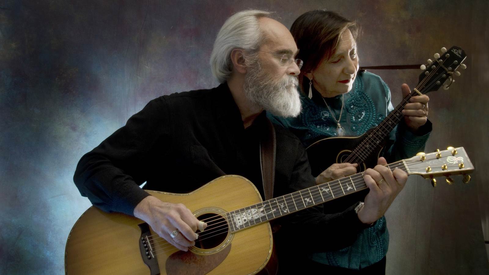 Two artists with their acoustic guitars looking to the right. One artist is wearing black and has a grey beard and hair. The other artist is behind him, she is wearing a denim jacket and playing the guitar.