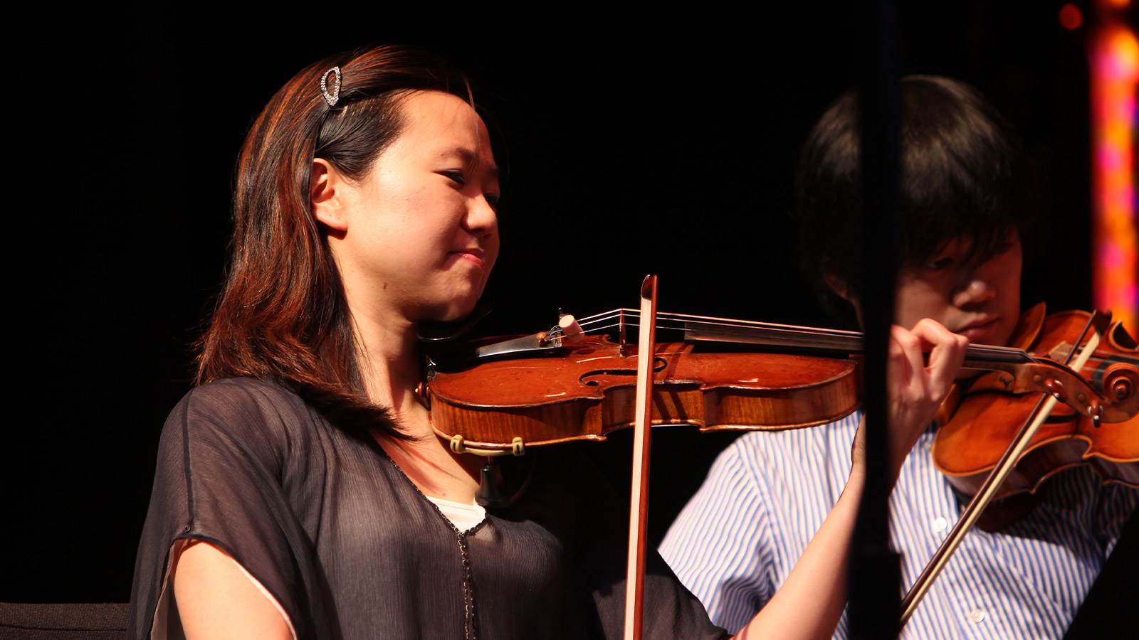 A violinist is playing her instrument. She has brown hair clipped back with a barrette in front. She is wearing a black top and is looking to the right. There is another violinist is behind her who is wearing a stripped button down. 