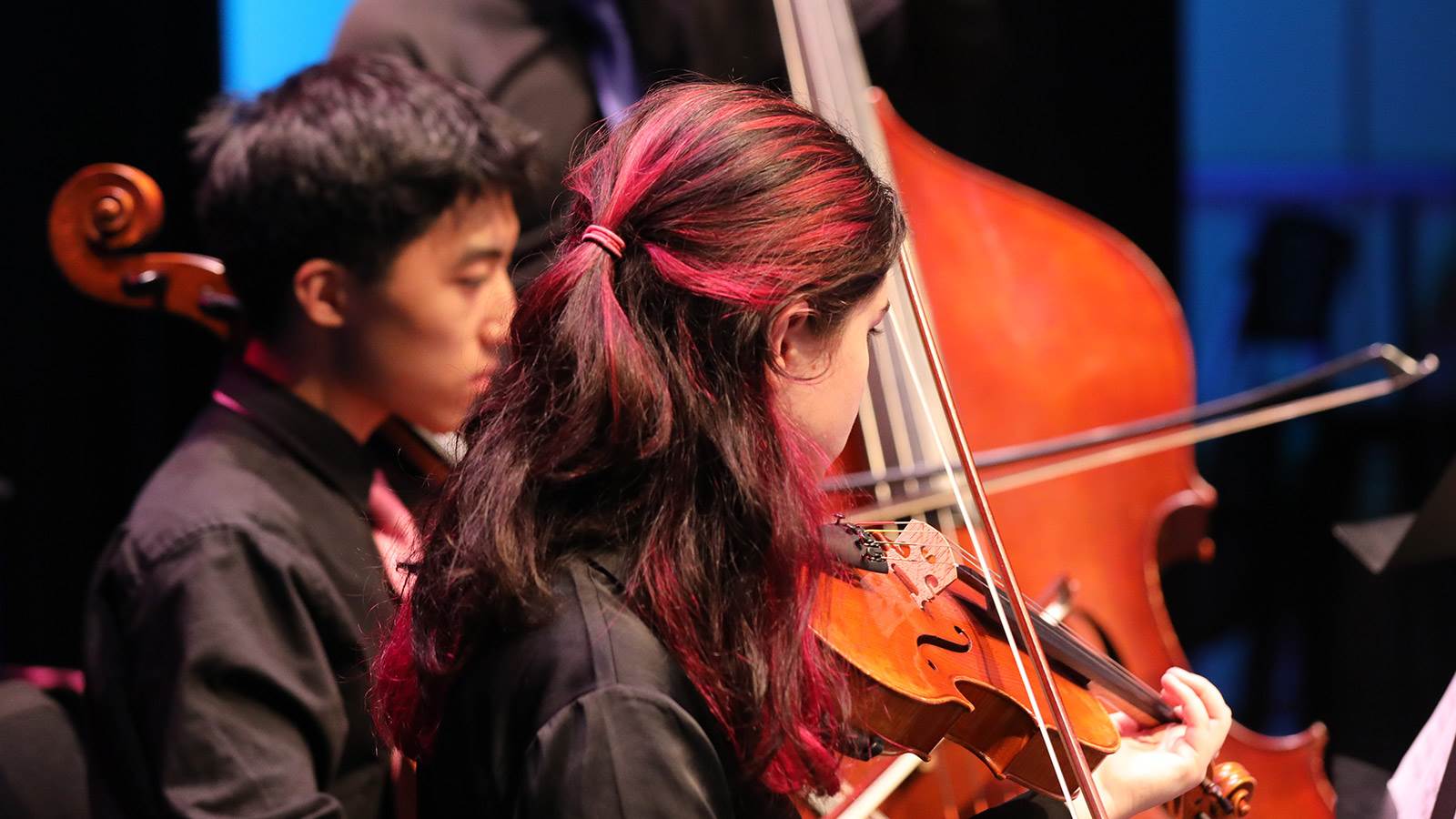 Young musicians playing their instruments. One the right is a young lady playing the violin and on her left is a young man playing a cello.