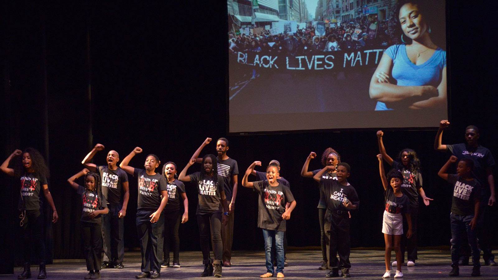 Many students standing on a stage, wearing black t-shirts. There is a projection in the background with a women crossing her arms and text next to her saying "Black Lives Matter"