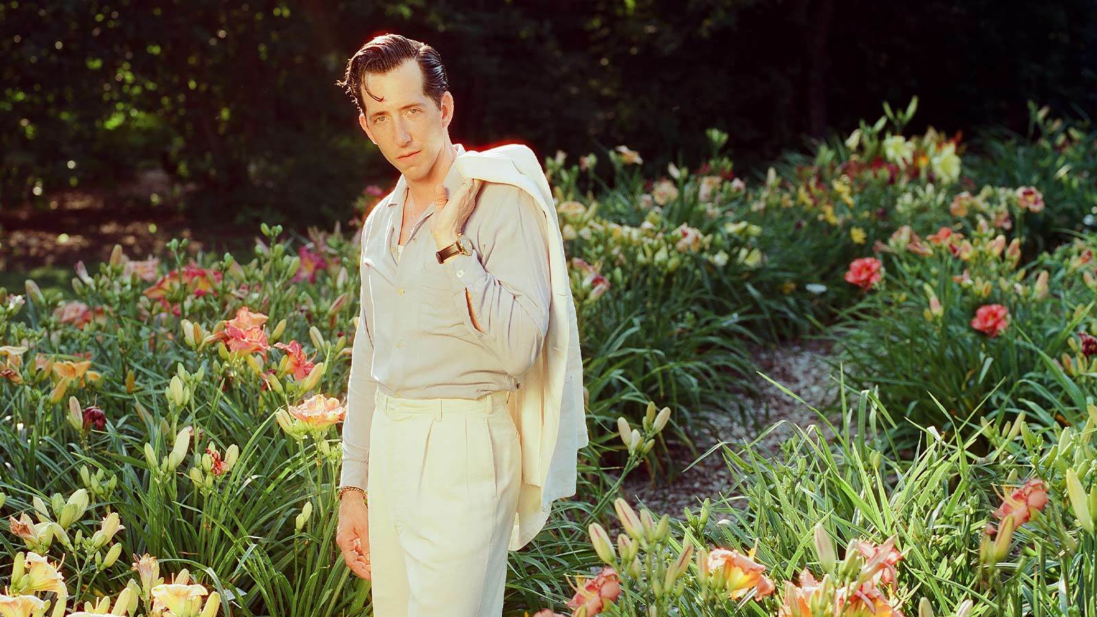 Artist Pokey Lafarge in a cream colored suit in front of a garden of lilies. He has is blazer draped over his shoulder.