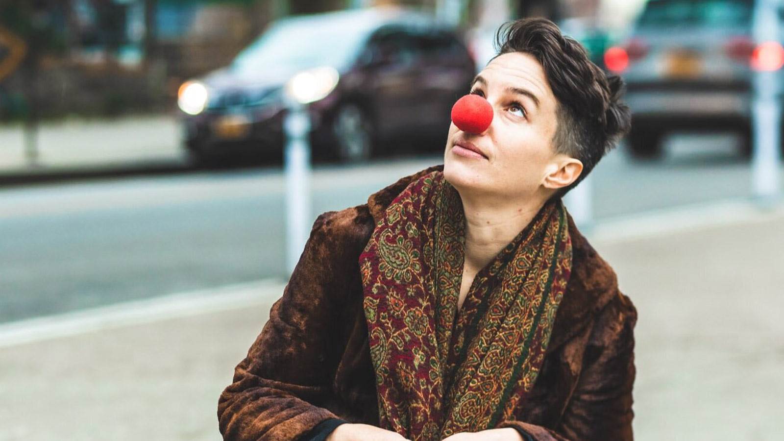 A person looking up at the sky, wearing a brown coat and a clown nose. They are near a city street.
