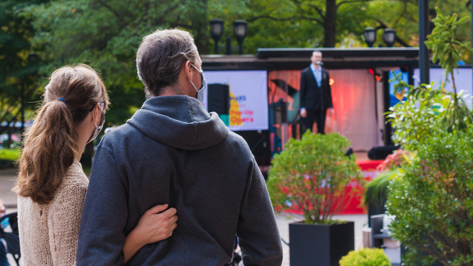 A woman and man watch a male singer perform from an outdoor stage on a truck in the daytime.
