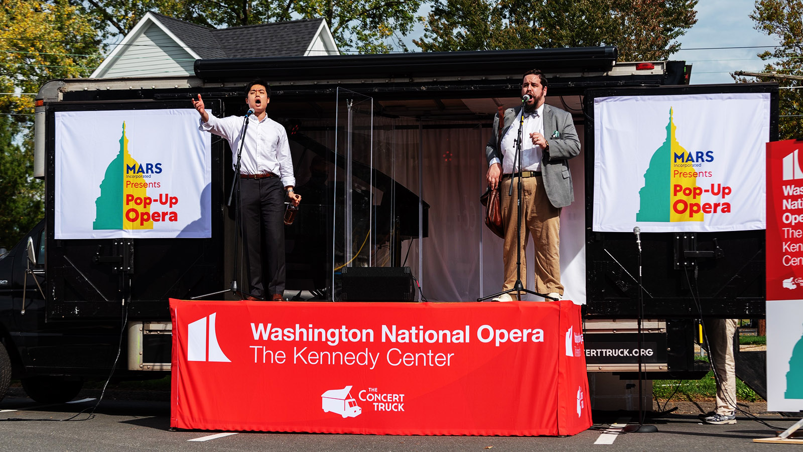 Two male singers perform from an outdoor stage on a truck in the daytime.